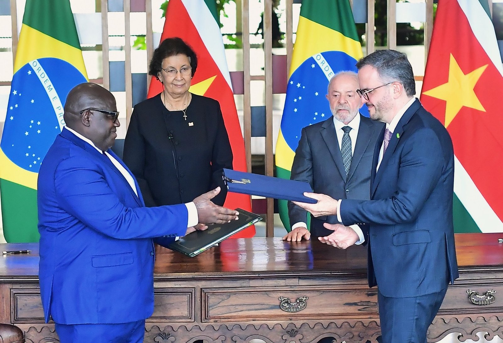 Fotografia em plano médio de uma cerimônia oficial de assinatura de acordos entre o Brasil e o Suriname. Em primeiro plano, dois homens de terno estão de perfil, um de frente para o outro, trocando pastas de documentos sobre uma grande mesa de madeira rústica e trabalhada.  À esquerda, um homem negro de terno azul-biciclete e óculos estende a mão direita para entregar uma pasta escura. À direita, um homem branco de terno azul-marinho e óculos segura uma pasta azul-escura.  Ao fundo, centralizados, estão mais duas autoridades observando o ato: uma mulher de blazer preto e colar discretamente sorrindo à esquerda, e o presidente brasileiro Luiz Inácio Lula da Silva à direita, vestindo terno cinza e gravata quadriculada. Atrás deles, quatro bandeiras oficiais estão hasteadas de forma intercalada: duas do Brasil (verde, amarela e azul) e duas do Suriname (faixas horizontais verdes, brancas e vermelha com uma estrela amarela no centro). A iluminação é clara e o ambiente é formal.