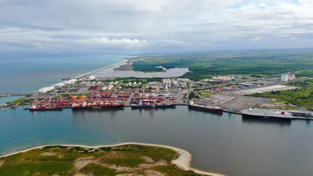 Fotografia aérea do Porto de Suape, em Pernambuco. A imagem apresenta navios atracados nos cais, áreas de movimentação de contêineres, tanques industriais e estruturas logísticas. Ao redor do complexo portuário, há áreas de vegetação, faixas de praia e o oceano ao fundo.
