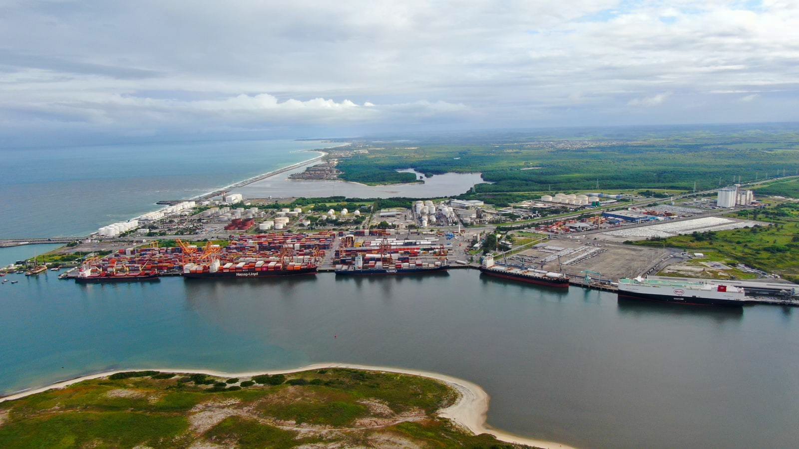 Fotografia aérea do Porto de Suape, em Pernambuco. A imagem apresenta navios atracados nos cais, áreas de movimentação de contêineres, tanques industriais e estruturas logísticas. Ao redor do complexo portuário, há áreas de vegetação, faixas de praia e o oceano ao fundo.