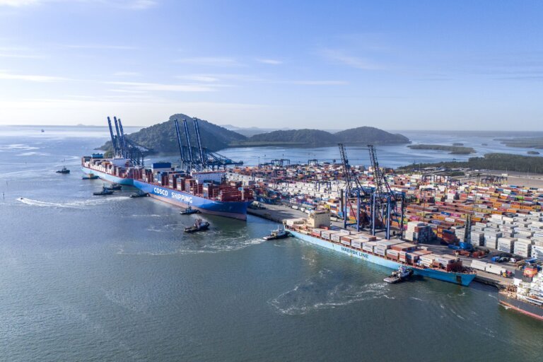 Uma vista aérea panorâmica e diurna mostra um movimentado terminal portuário de contêineres à beira-mar, cercado por águas calmas e colinas verdes ao fundo sob um céu claro.