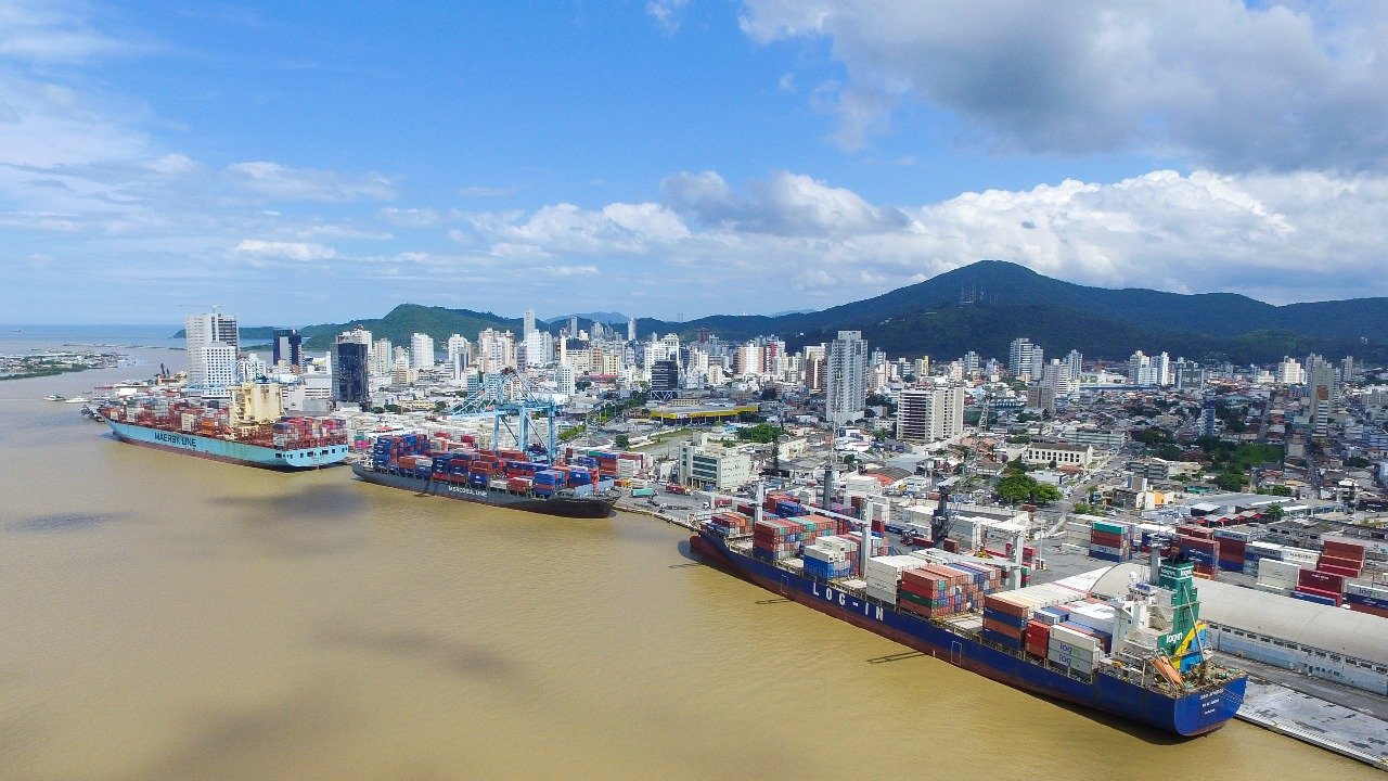 Vista aérea do Porto de Itajaí, em Santa Catarina. Três navios cargueiros atracados aparecem ao lado de pilhas de contêineres coloridos e estruturas portuárias. Ao fundo, a área urbana da cidade, com muitos edifícios, é cercada por morros cobertos por vegetação. O céu azul com nuvens completa a paisagem.