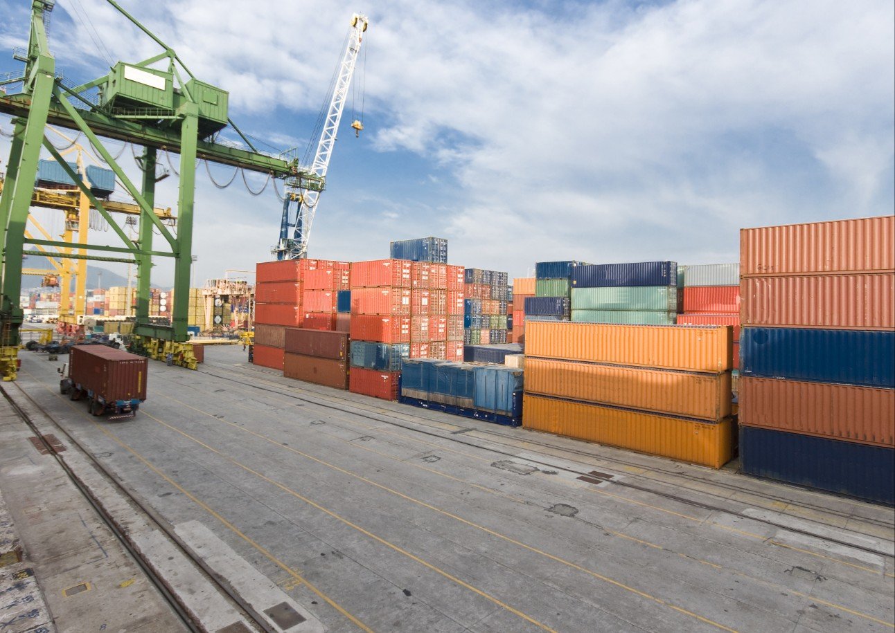 Pátio de terminal portuário com contêineres empilhados e guindastes para movimentação de cargas.