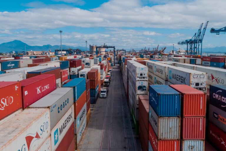 Imagem mostra uma área portuária com pilhas de contêineres coloridos organizados em fileiras. Ao centro, há uma via interna com caminhões circulando entre as cargas. Ao fundo, aparecem guindastes, estruturas operacionais do porto, montanhas e céu parcialmente nublado.