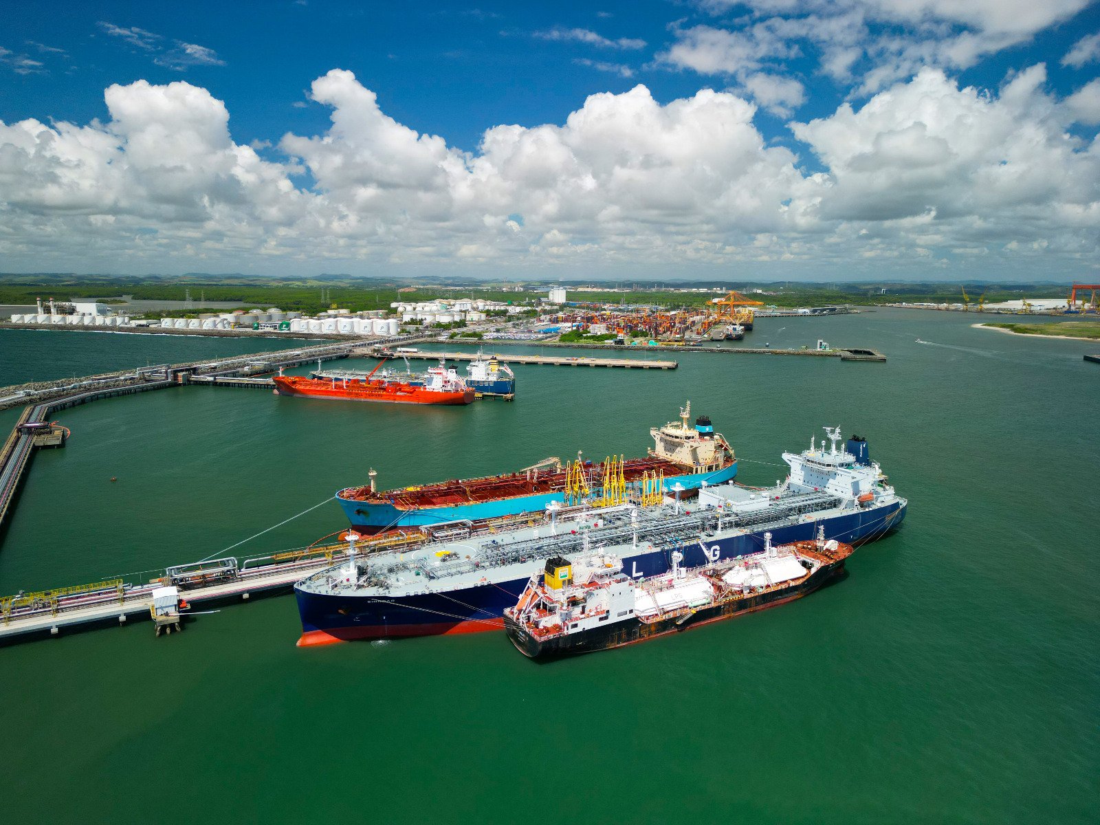 Imagem aérea do Complexo Industrial Portuário de Suape, em Pernambuco. A foto mostra navios atracados em píeres de operação, com estruturas de dutos e equipamentos portuários sobre a água. Ao fundo, aparecem tanques de armazenagem, áreas operacionais, guindastes, terminal de contêineres e a paisagem do entorno, sob céu azul com nuvens.
