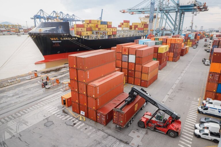 Imagem mostra um terminal portuário de contêineres, com navio atracado ao cais e pilhas de contêineres coloridos organizadas no pátio. Há guindastes portuários, caminhões, veículos de apoio e equipamento de movimentação de contêiner em operação na área do terminal.