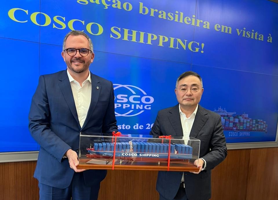 Dois homens de terno preto seguram maquete de navio de carga.