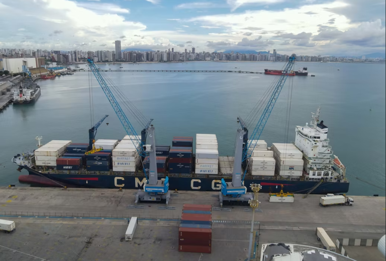 Vista aérea de um navio cargueiro atracado no Porto de Fortaleza, com contêineres empilhados e dois guindastes realizando a operação de carga e descarga. Ao fundo, aparecem o mar, outras embarcações e a orla urbana da cidade sob um céu parcialmente nublado.