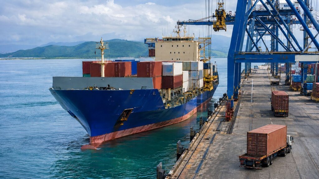 Navio porta-contêineres atracado em terminal portuário, com guindastes e caminhões transportando cargas.