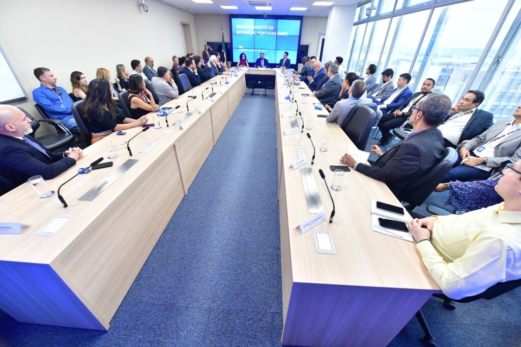 na imagem, foto da reunião de apresentação do grupo de trabalho. Pessoas estão sentadas em uma mesa retangular e há tres pessoas ao centro que conduzem a apresentação.