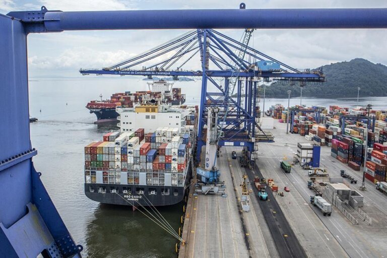 Vista aérea de terminal portuário, com navio porta-contêineres atracado, guindastes no cais e contêineres organizados na área operacional.
