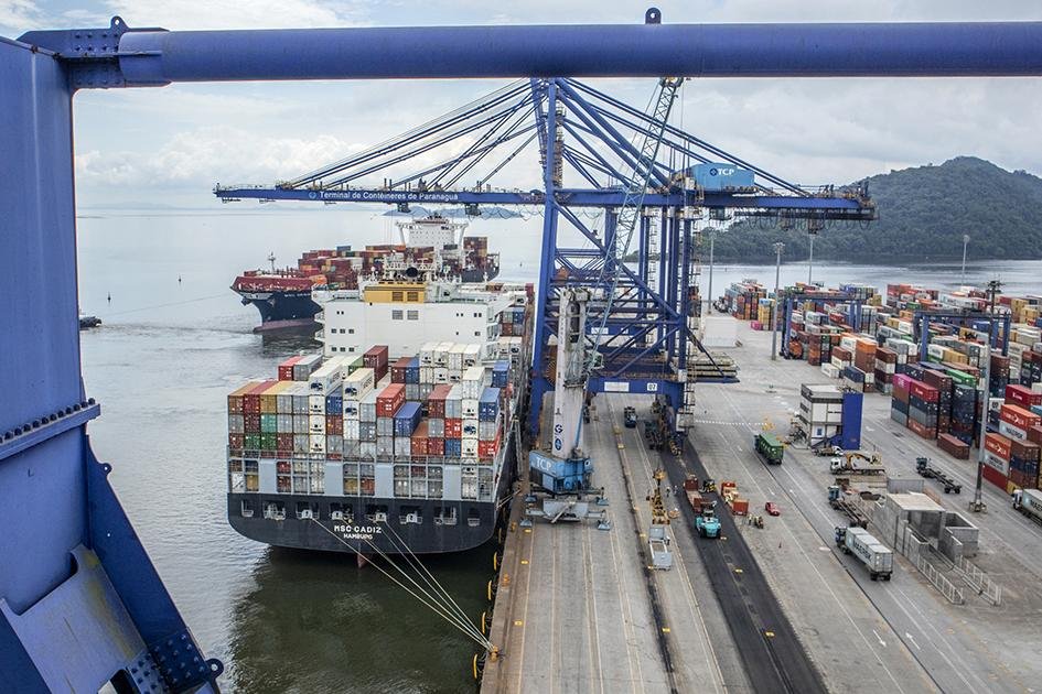 Vista aérea de terminal portuário, com navio porta-contêineres atracado, guindastes no cais e contêineres organizados na área operacional.