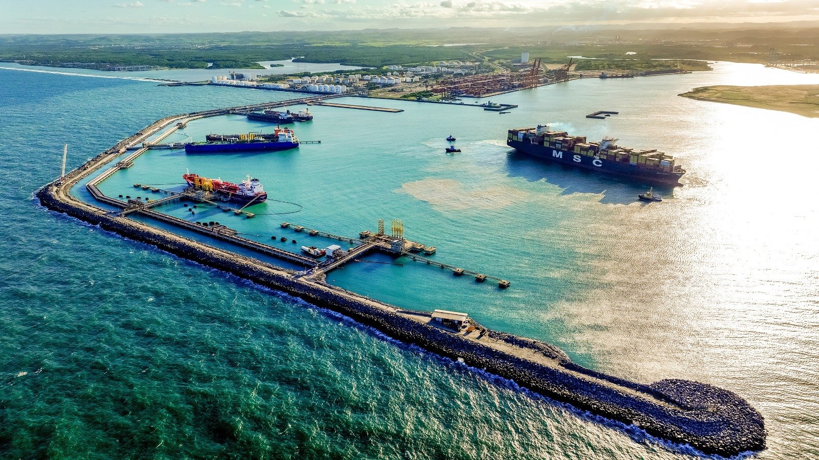 Vista aérea do Porto de Suape, em Pernambuco, com navios atracados e em operação, incluindo um porta-contêineres e embarcações para granéis líquidos. A imagem mostra píeres, estruturas portuárias, áreas de armazenagem e o canal de acesso cercado pelo mar.
