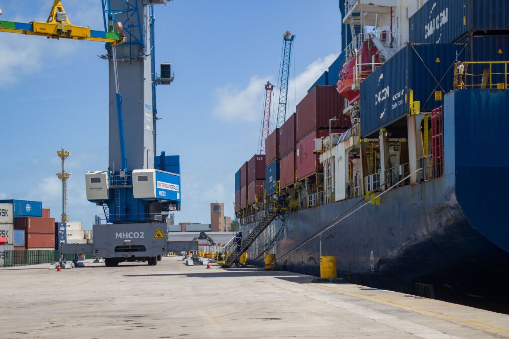 Navio porta-contêineres atracado em terminal portuário, com guindastes e contêineres durante operação de carga e descarga.
