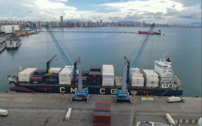 Vista aérea de um navio cargueiro atracado no Porto de Fortaleza, com contêineres empilhados e dois guindastes realizando a operação de carga e descarga. Ao fundo, aparecem o mar, outras embarcações e a orla urbana da cidade sob um céu parcialmente nublado.